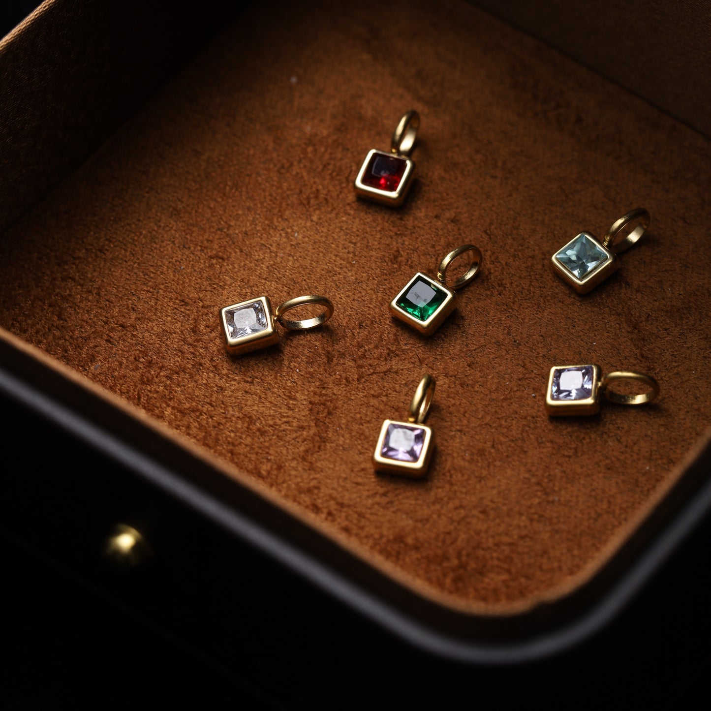A collection of square-cut birthstone charms (January - Garnet, February - Amethyst) in gold-plated settings, laid in a velvet jewelry box.